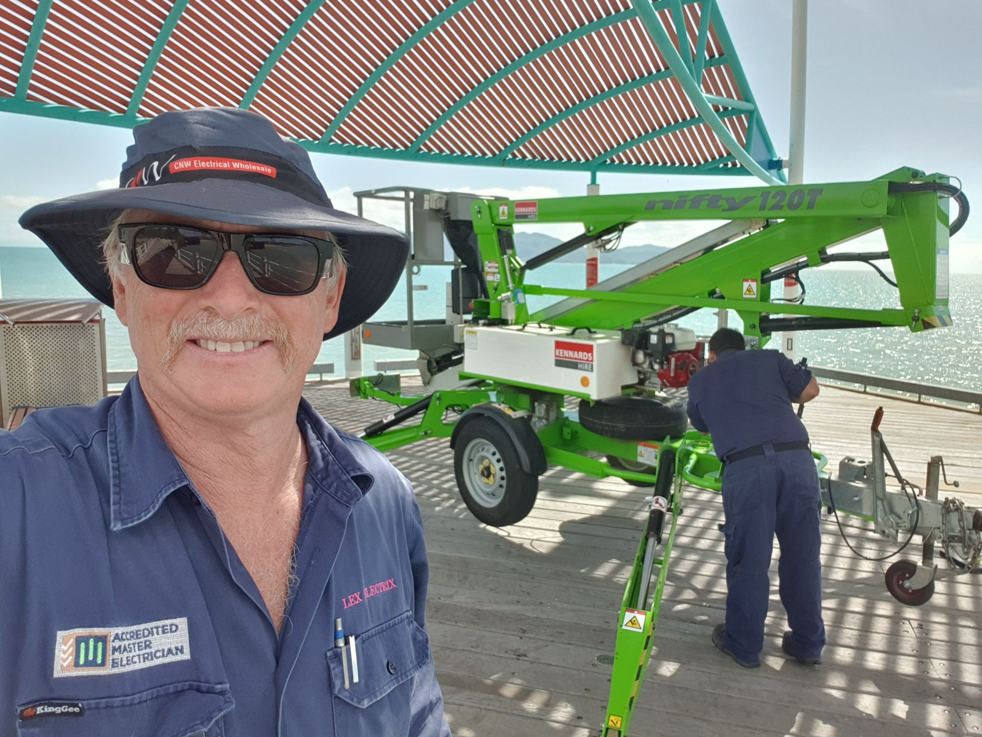 A Man Wearing A Hat And Sunglasses Smiling At the Camera With A Man Working On A Trailer Behind Him— Lex Electrix In West End, QLD
