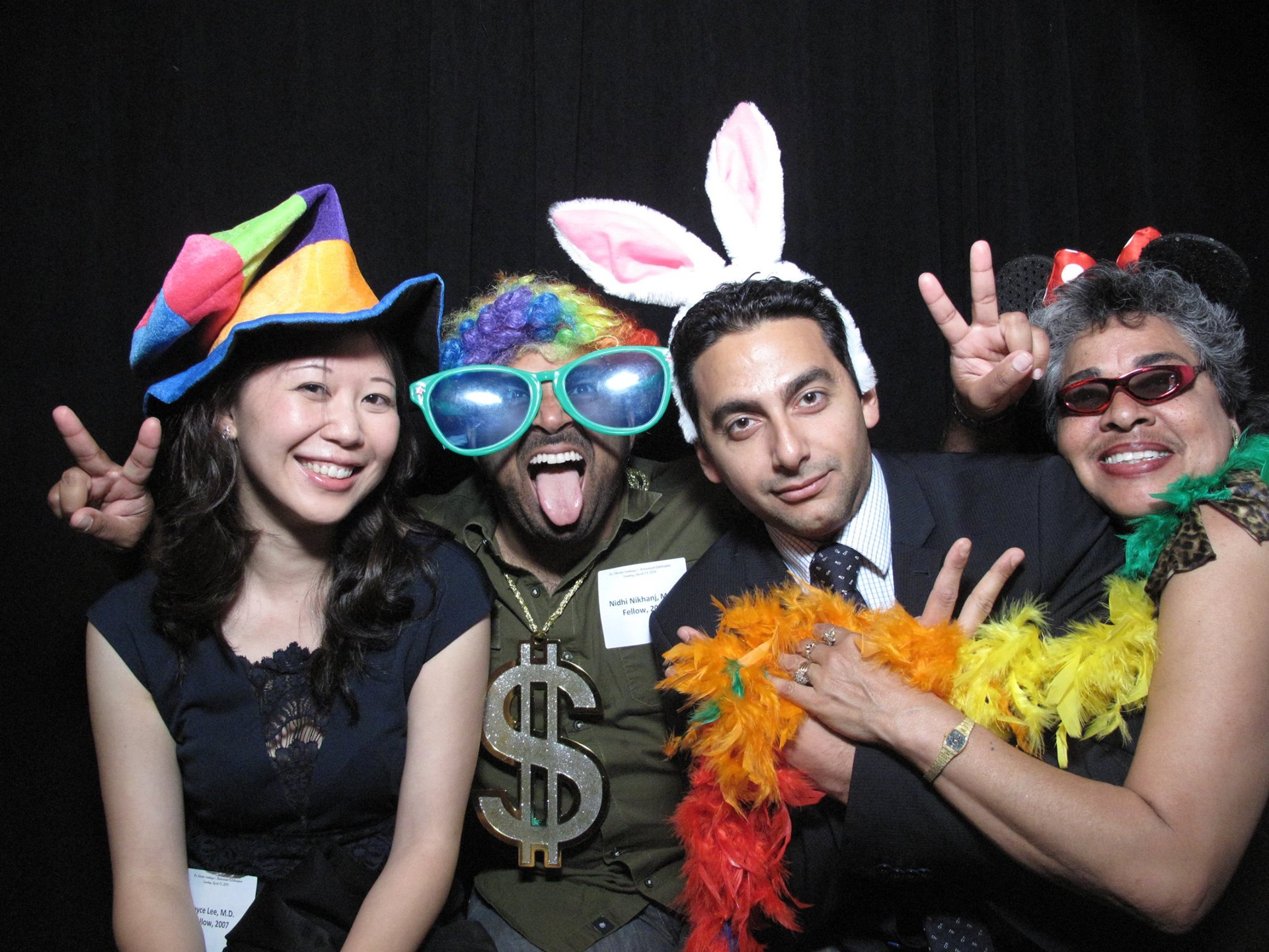 Four people in colorful costumes posing playfully at a party, smiling and making peace signs.