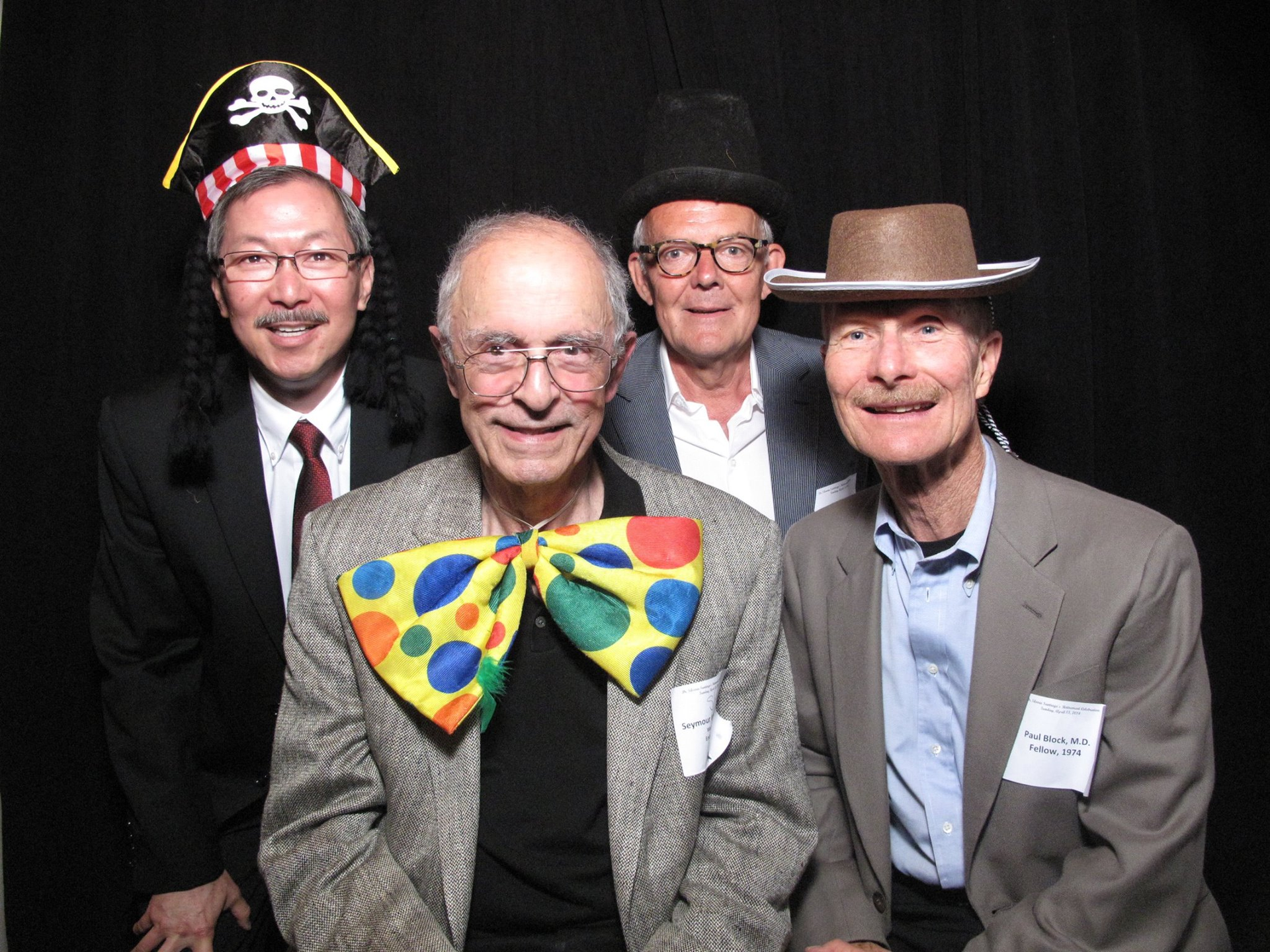 Four men in suits and hats posing together, one with a colorful clown bow tie.