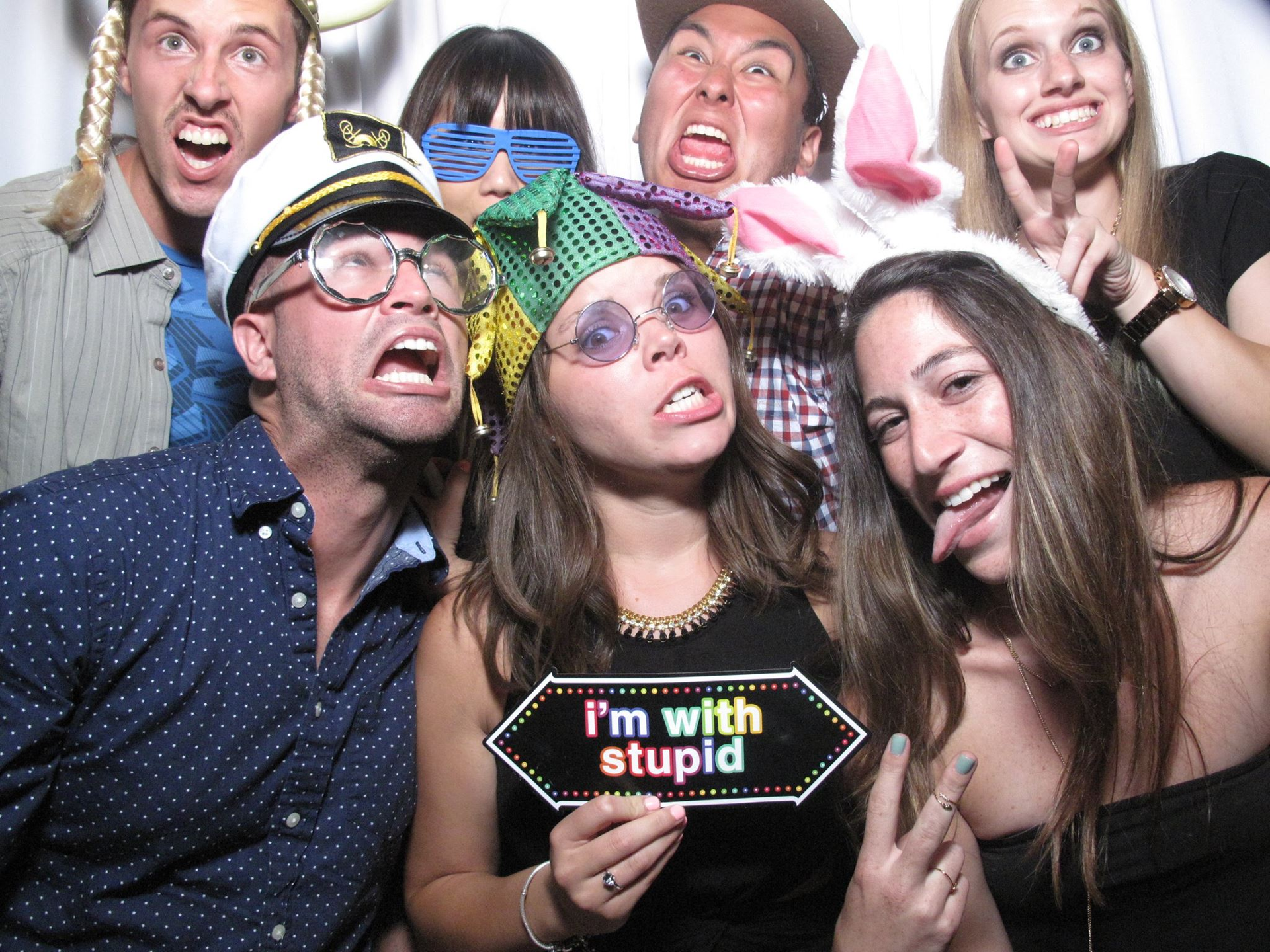 Group of people in silly hats and glasses posing for a photo, holding a sign that says “I’m with stupid”