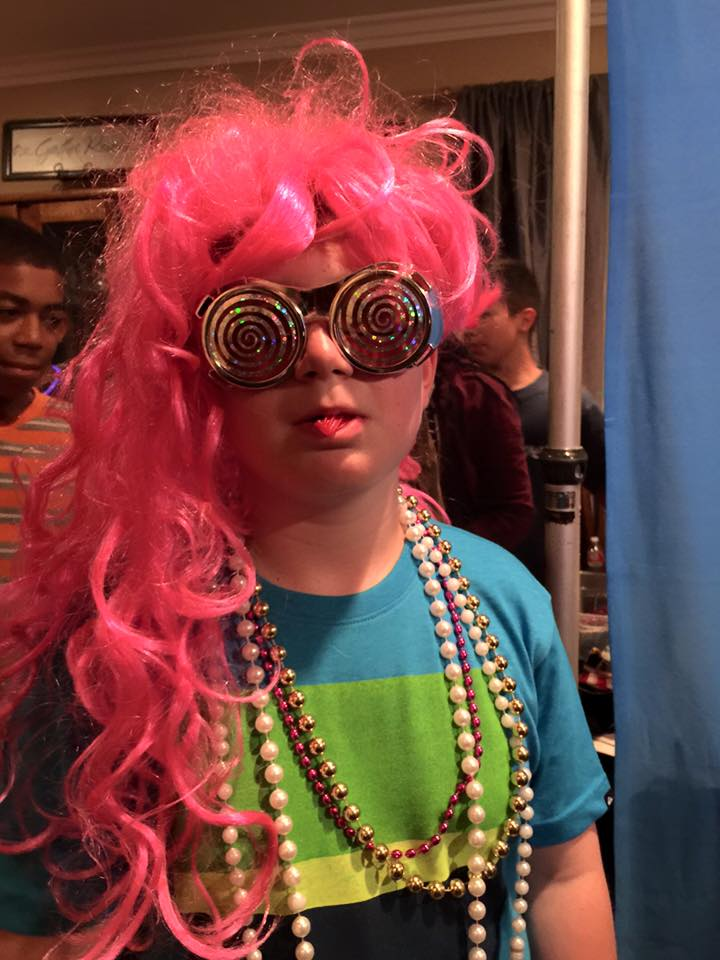 Child in bright pink wig and spiral glasses, wearing colorful beads and a blue-green shirt indoors.