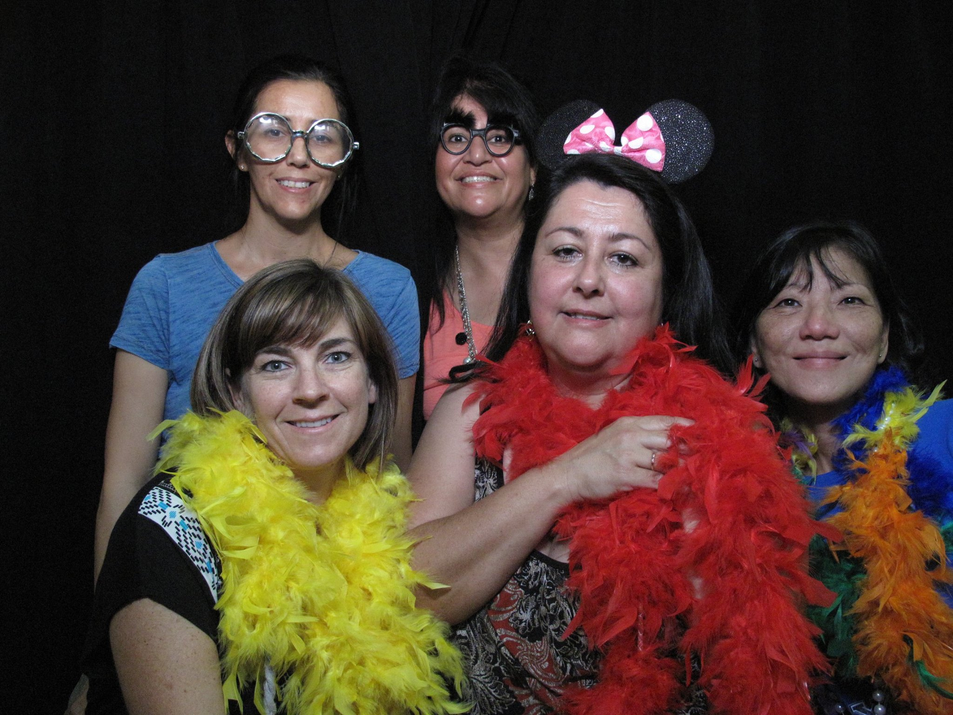 Five people pose together against a dark background, wearing colorful feather boas and playful accessories.