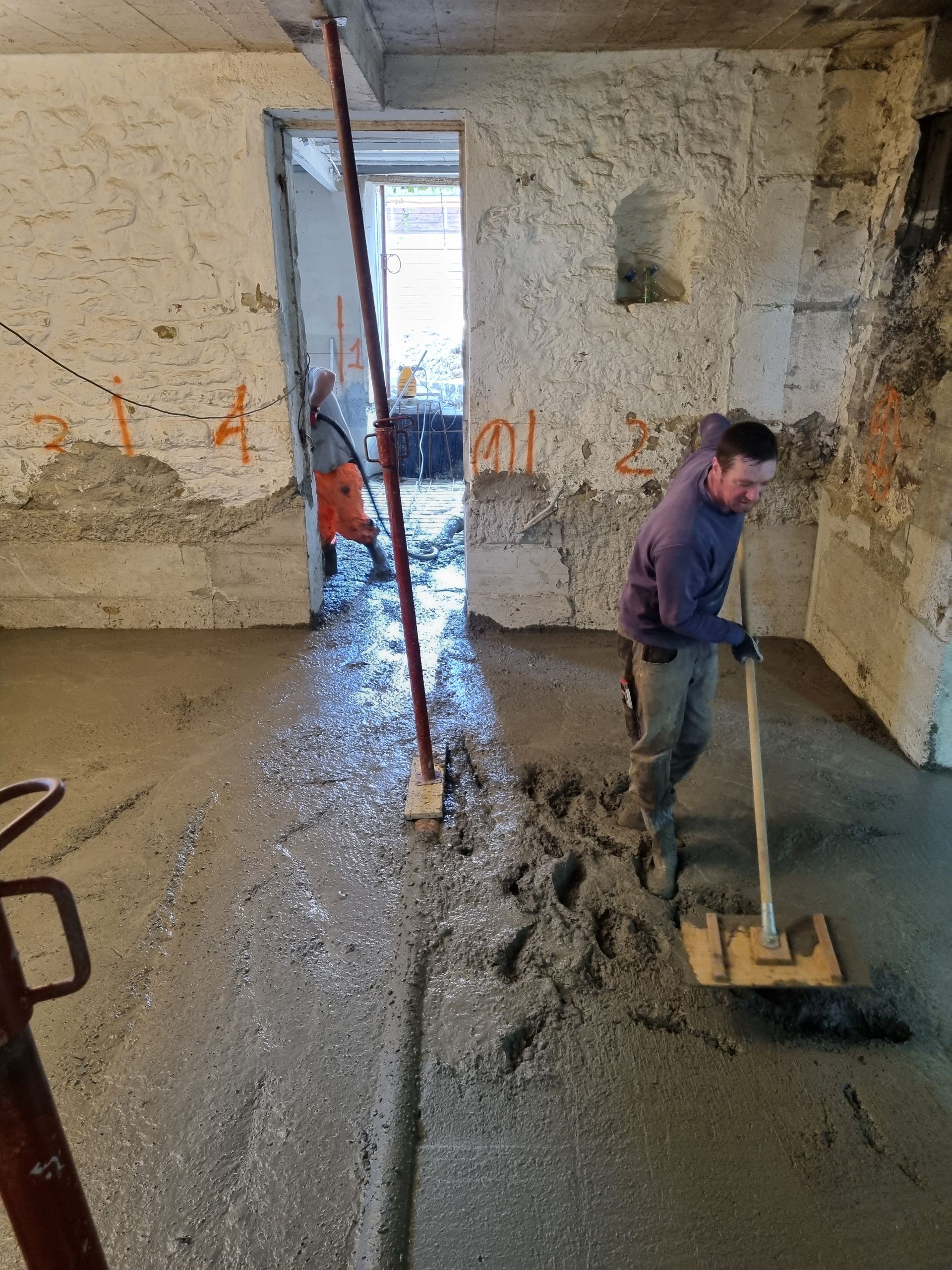 Un ouvrier vêtu d'une chemise violette utilise un outil à long manche pour niveler le béton frais sur le sol d'un sous-sol en cours de construction.