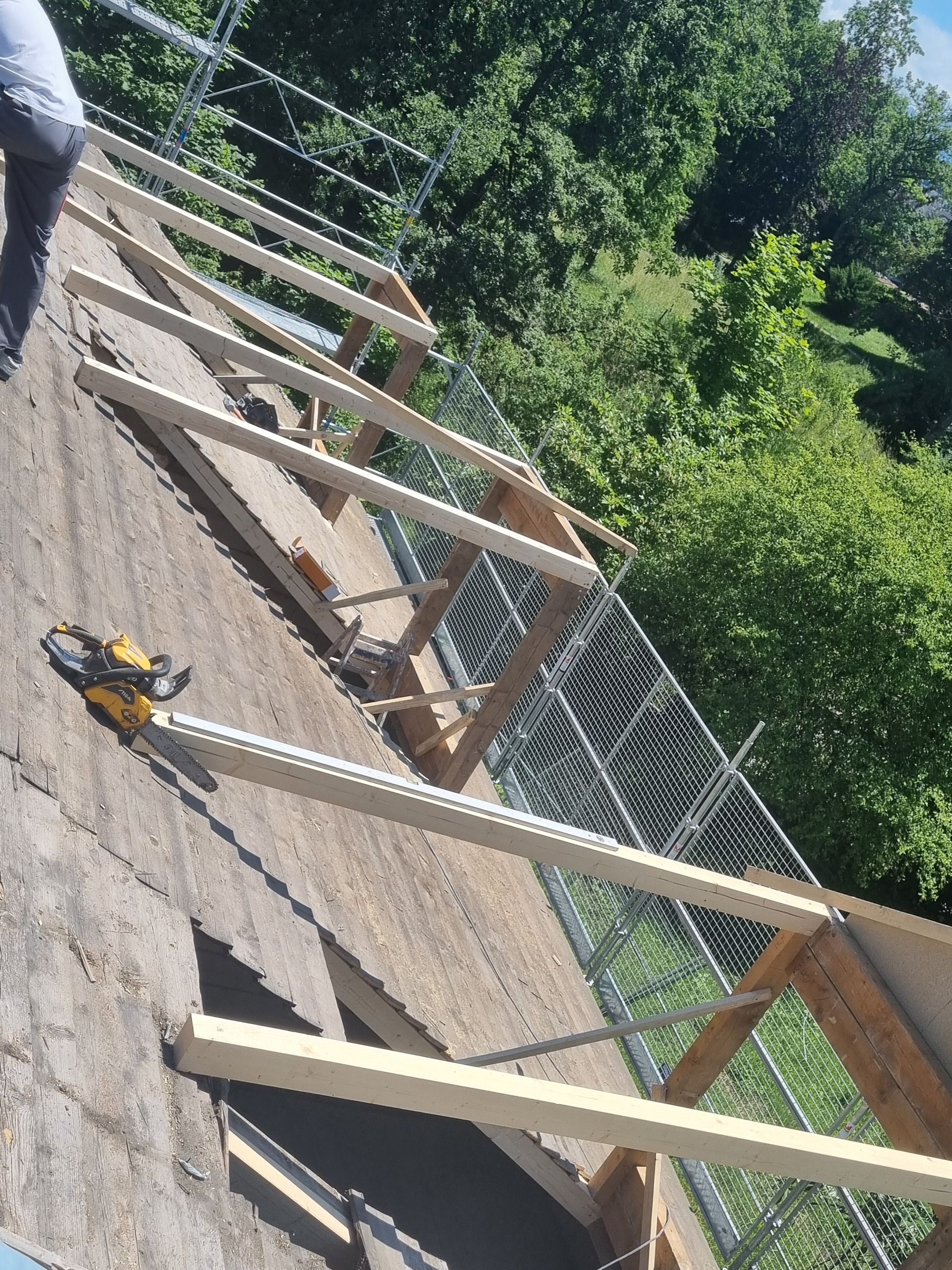 Une personne marche sur un toit en pente en construction, avec une charpente en bois, une tronçonneuse et une clôture métallique en arrière-plan.