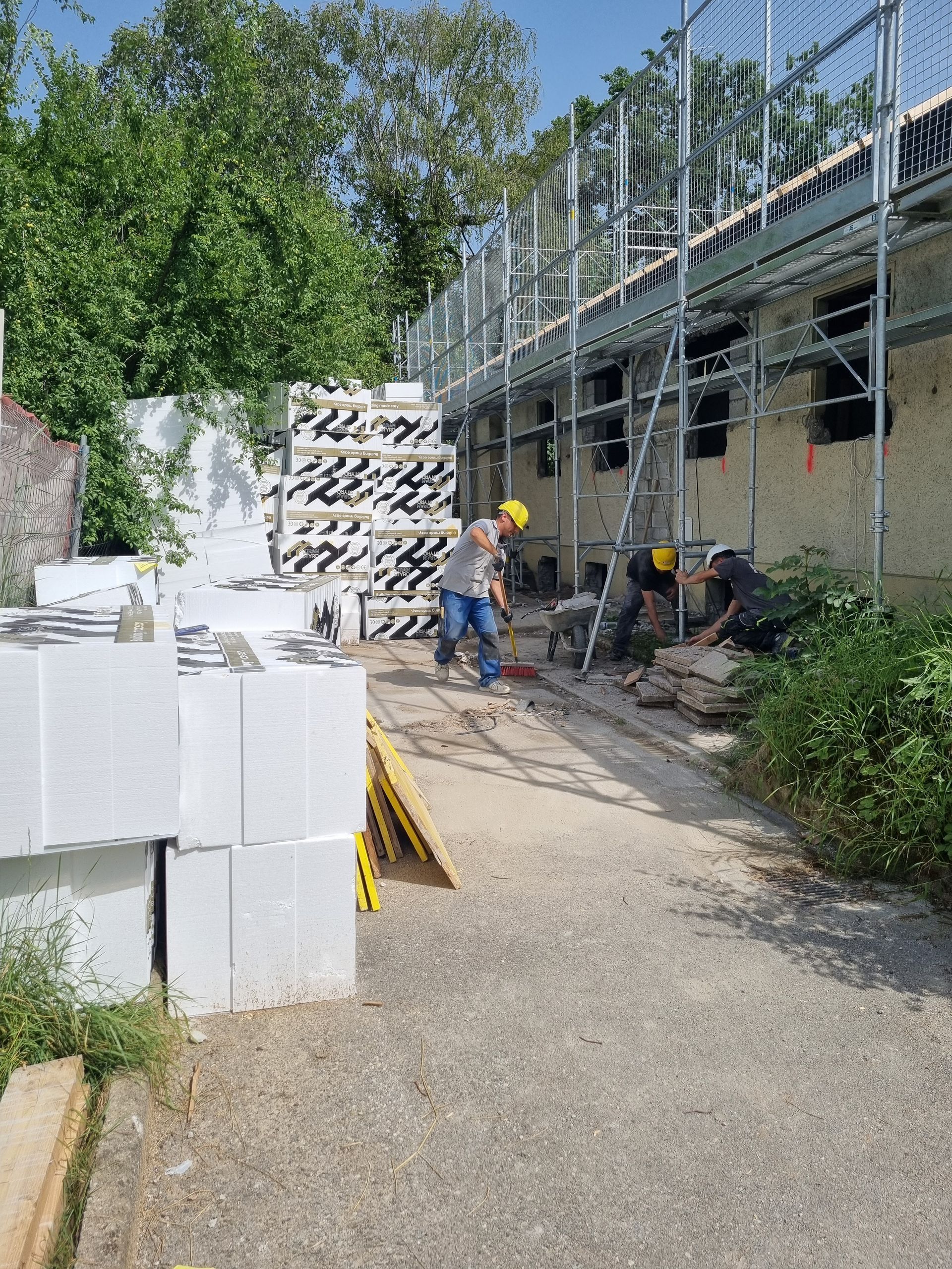 Des ouvriers du bâtiment, équipés de tenues de sécurité, travaillent à l'extérieur d'un bâtiment avec des échafaudages et des blocs d'isolation blancs empilés.