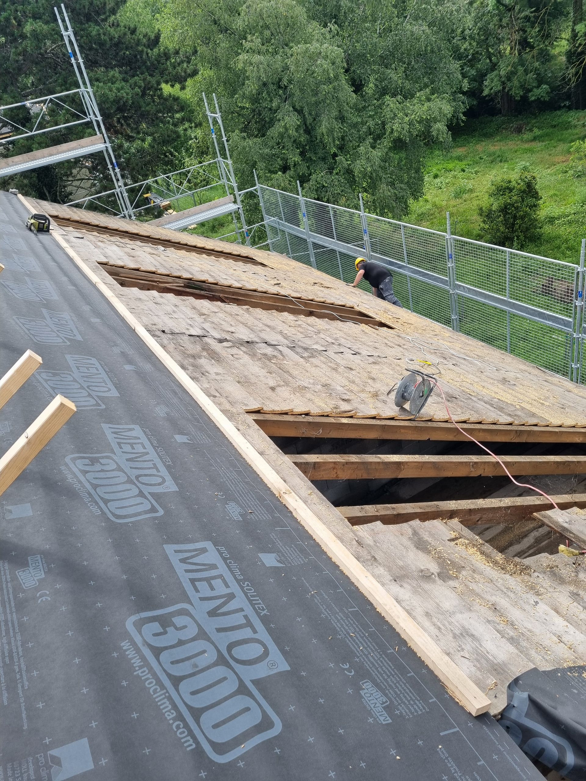 Un ouvrier répare le toit d'une maison avec ses chevrons en bois apparents et sa sous-couche noire, vu d'en haut près d'un échafaudage.