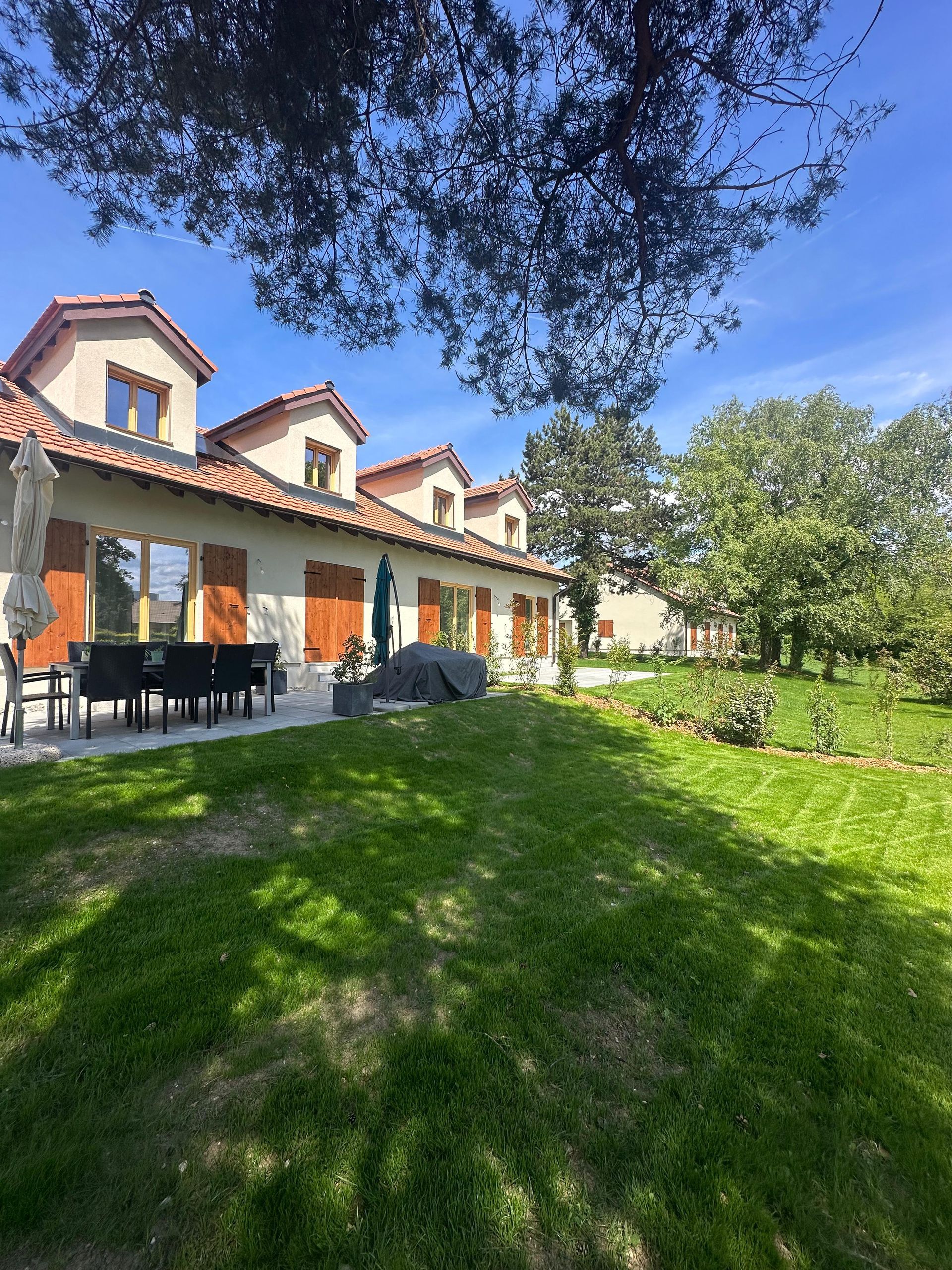 Une maison claire avec un toit de tuiles rouges, des lucarnes et une terrasse, le tout se détachant sur une pelouse verte et un ciel bleu azur.
