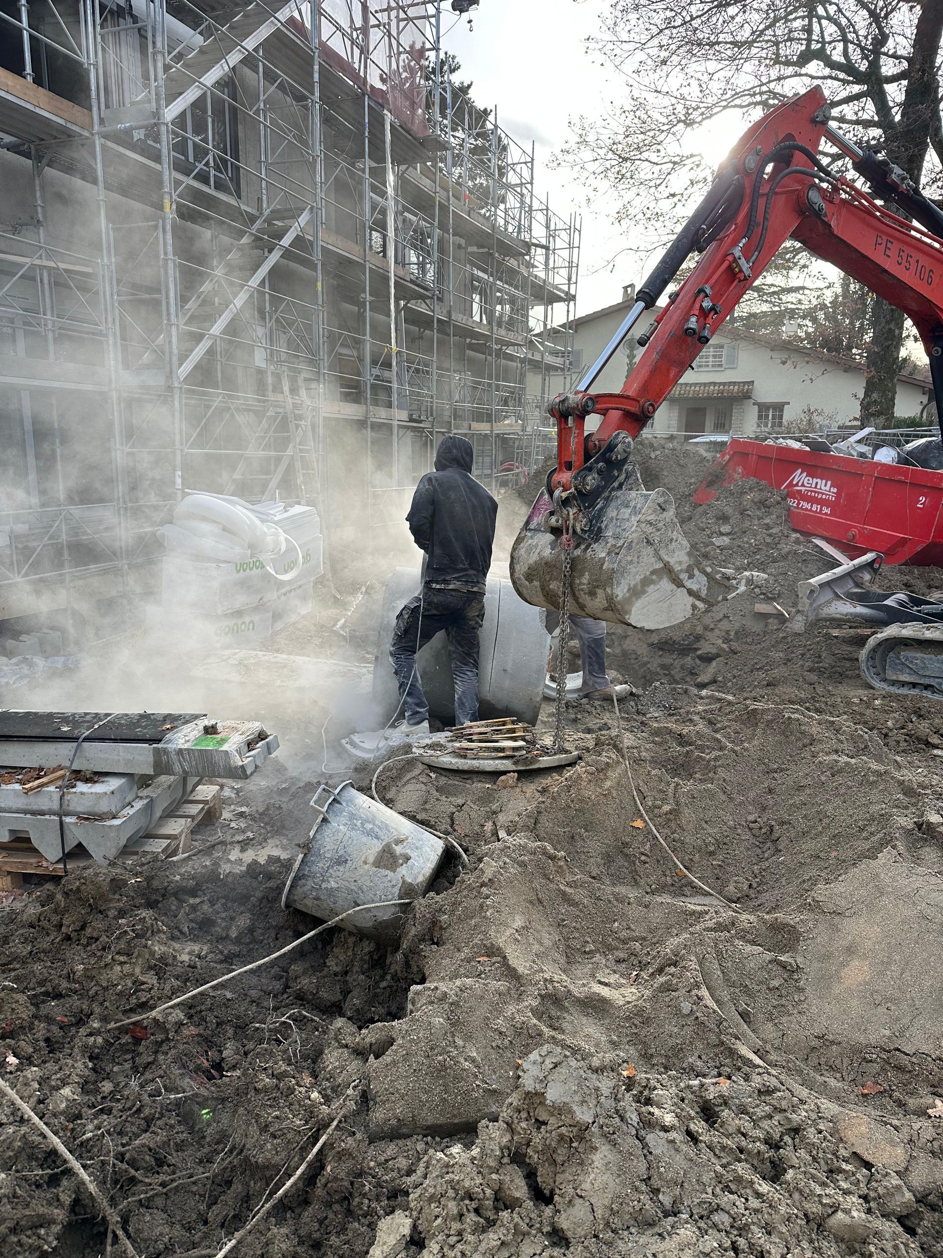 Un ouvrier utilise un outil manuel sur un tuyau en béton près d'une excavatrice rouge sur un chantier boueux avec des échafaudages à proximité.