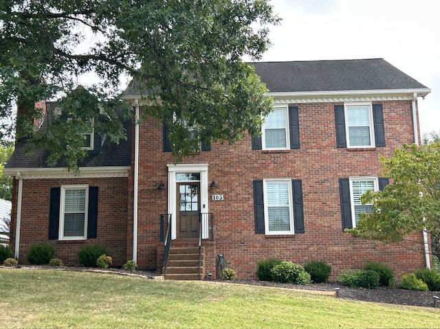 Brick two-story house with black shutters, a dark roof, and a small front yard with bushes.