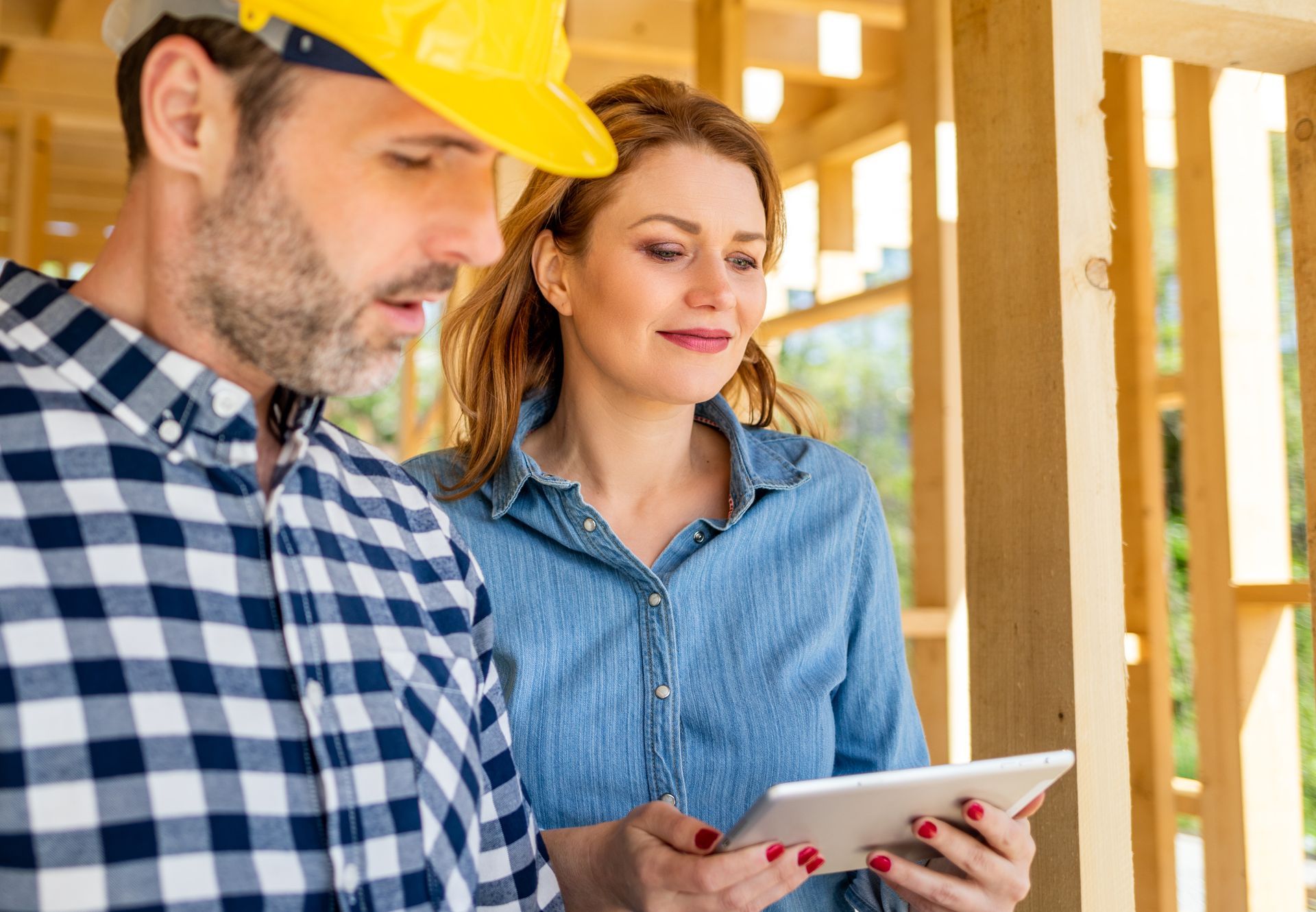 Man in hard hat and woman looking at a tablet inside a wooden building frame.