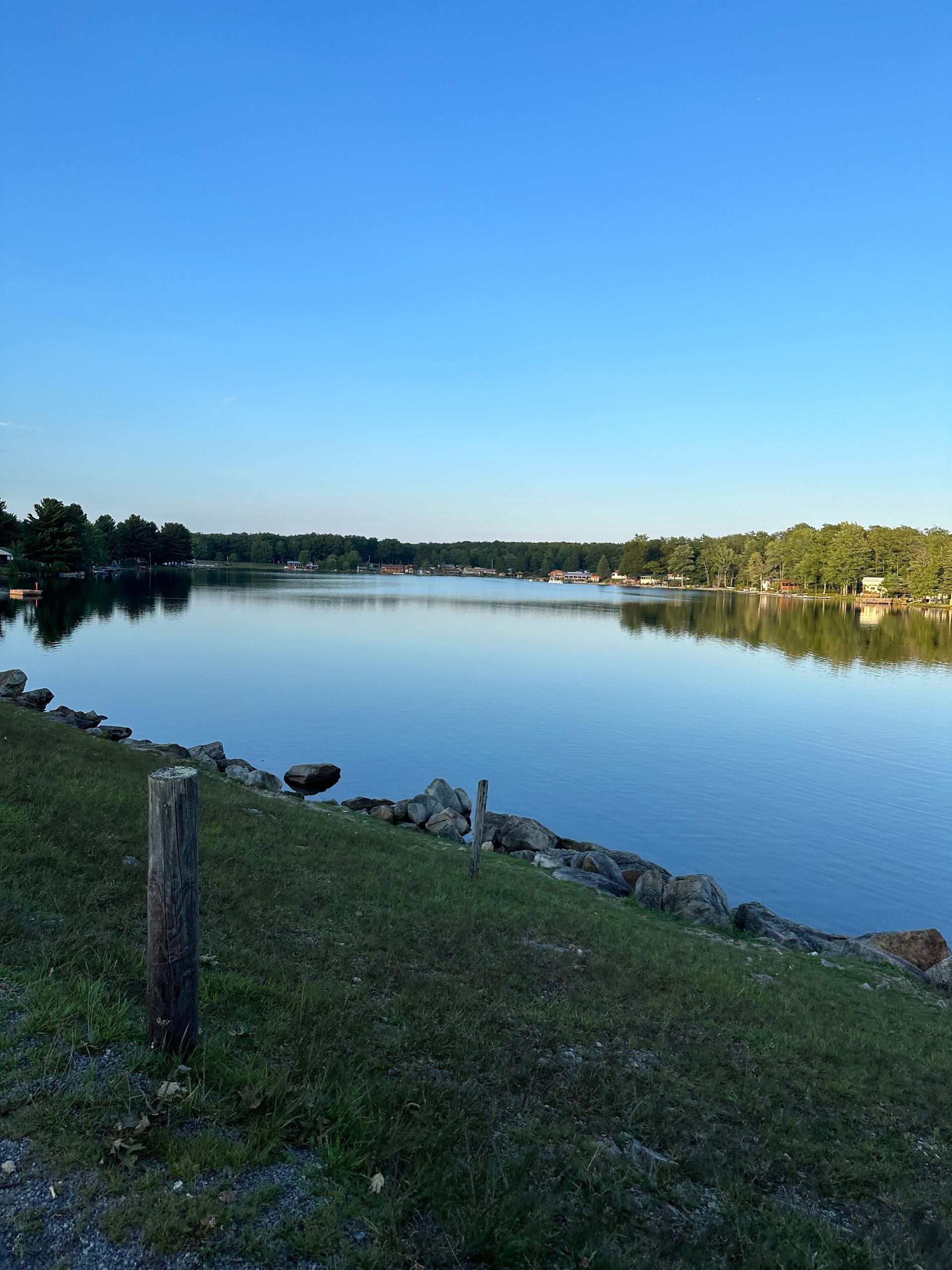 A calm lake reflects a clear blue sky, bordered by a rocky shoreline and a grassy slope with a lone wooden post.