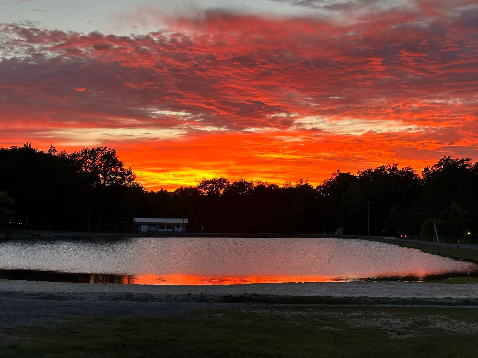 A vibrant sunset with fiery red and orange clouds reflecting over a calm lake bordered by a dark forest silhouette.