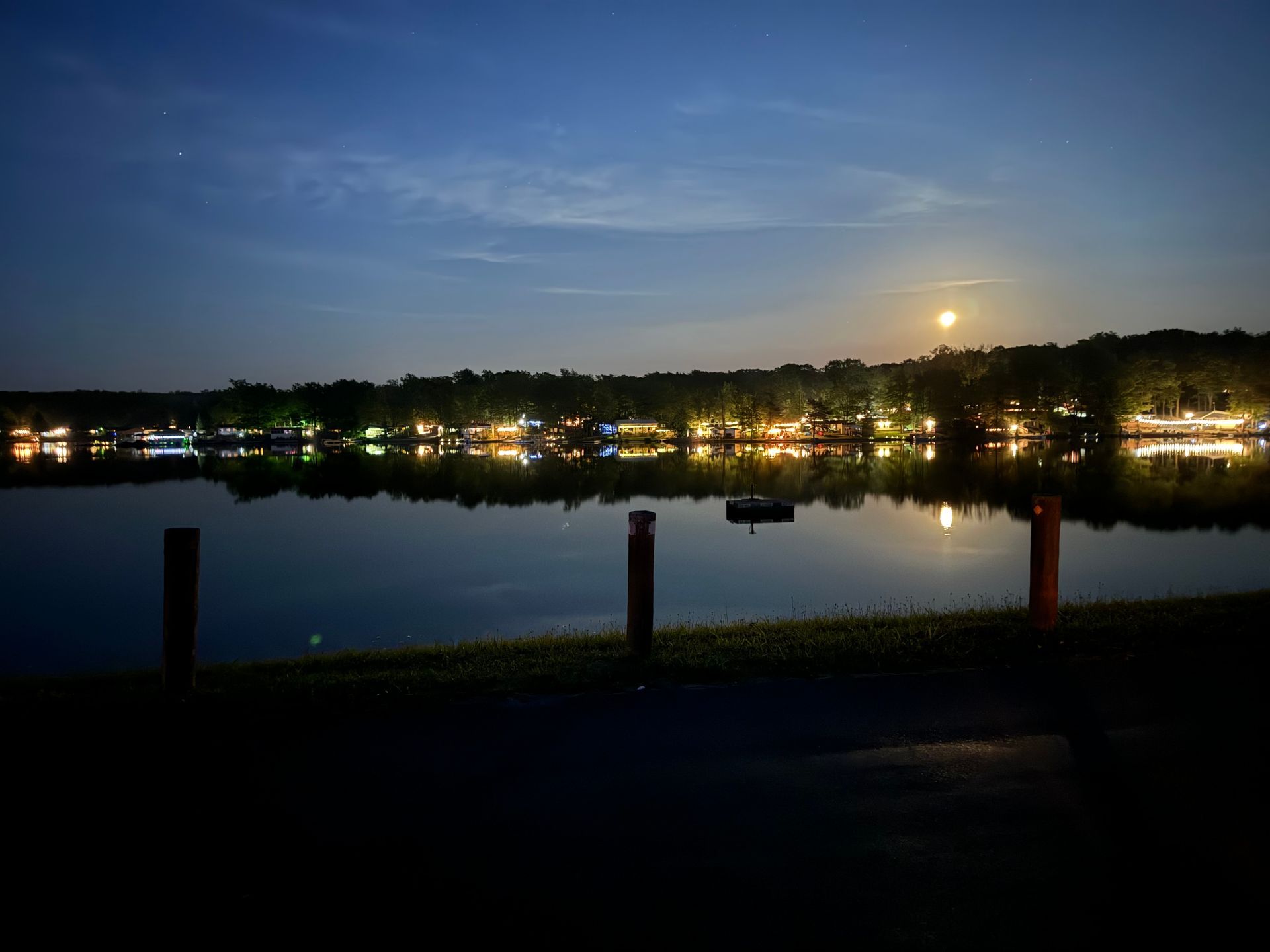 A calm lake reflects a line of glowing shore lights and a bright moon in the evening sky, viewed past three wooden posts.