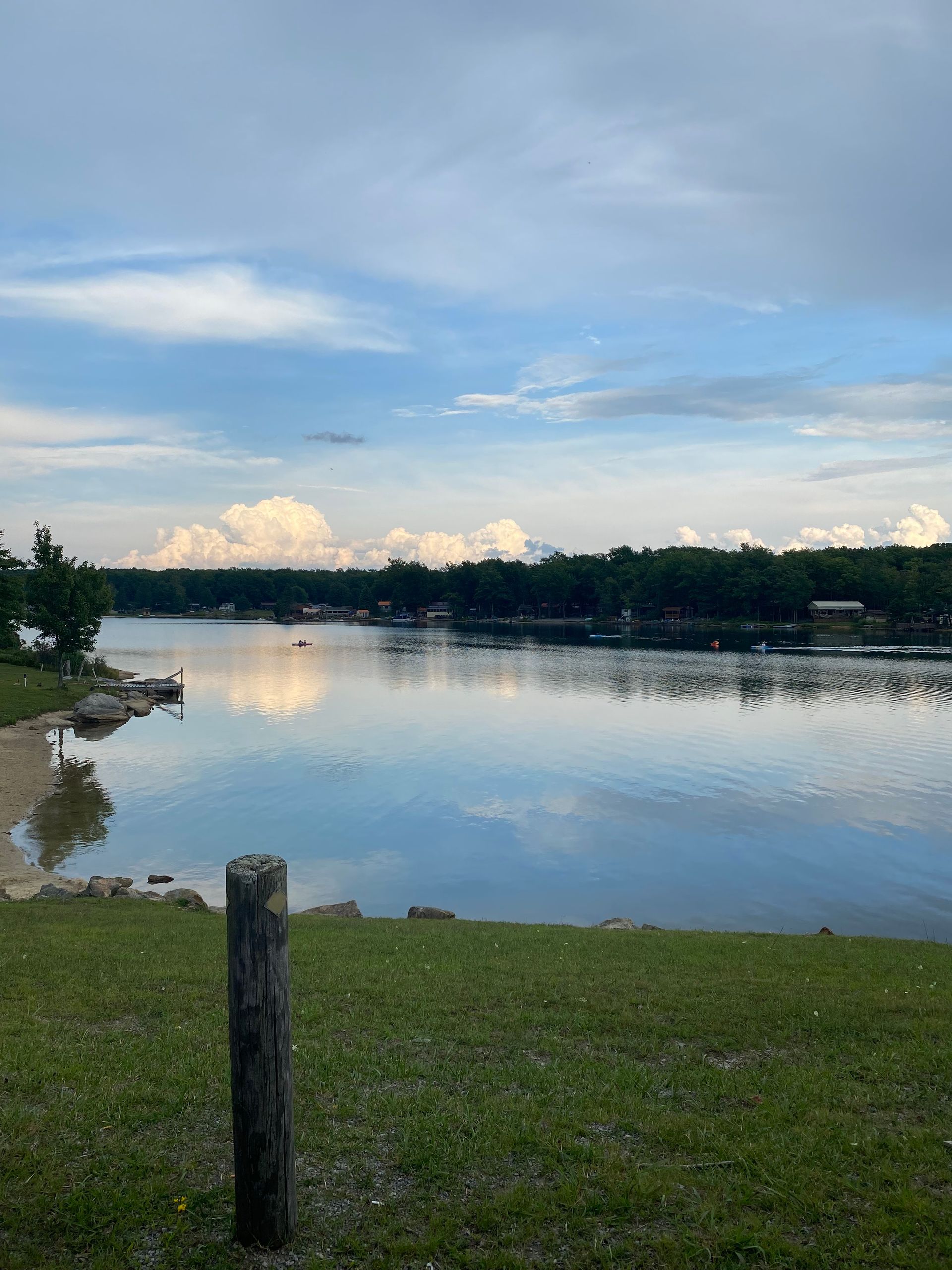 A tranquil lake under a blue sky with white clouds, featuring a grassy foreground and a wooden post.