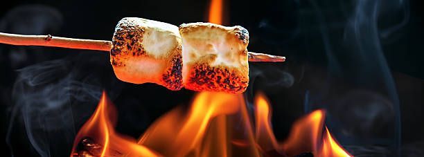 A toasted marshmallow on a stick held over an open campfire flame, with visible wisps of smoke against a dark background.