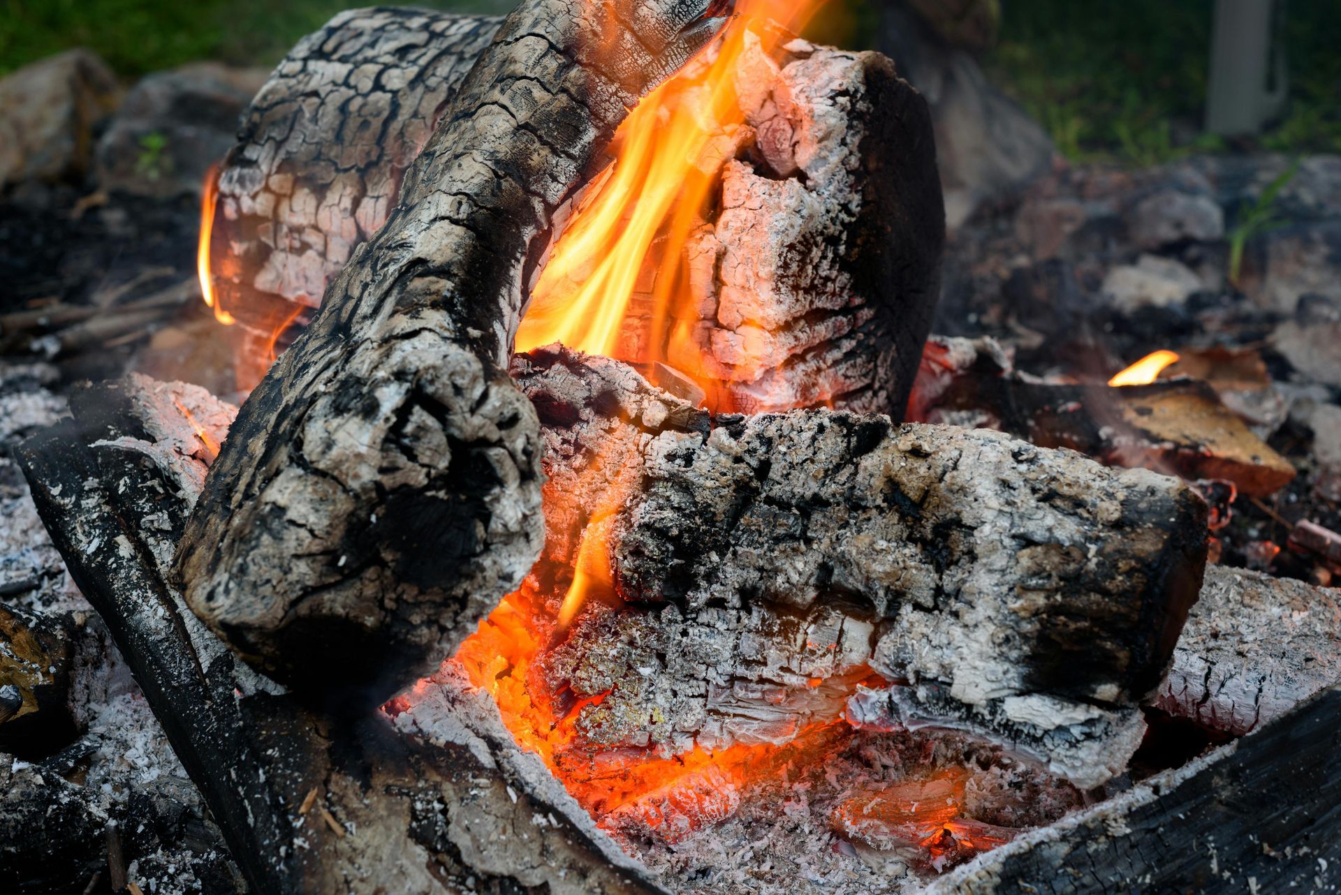 Bright orange flames flicker between charred, gray logs in a rustic outdoor fire pit.