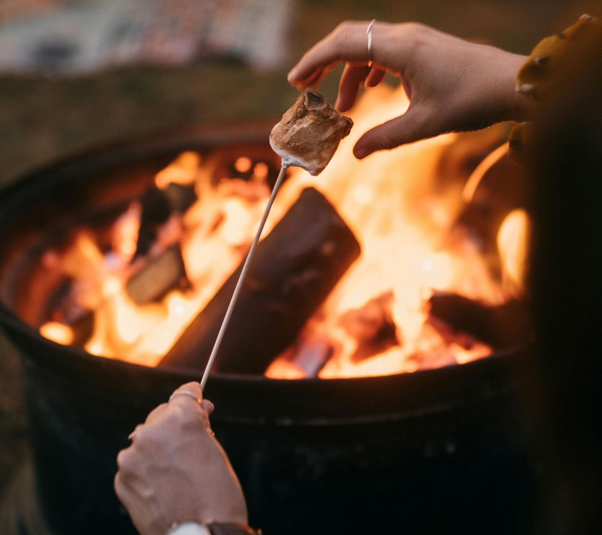 A hand toasts a golden-brown marshmallow on a metal skewer over a glowing campfire.