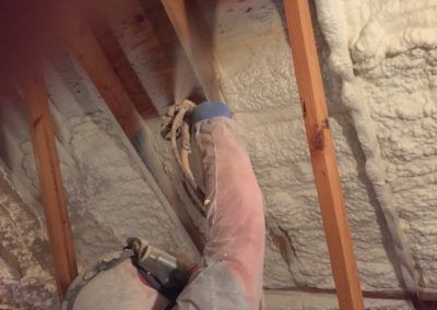 A person is spraying foam on the ceiling of an attic.