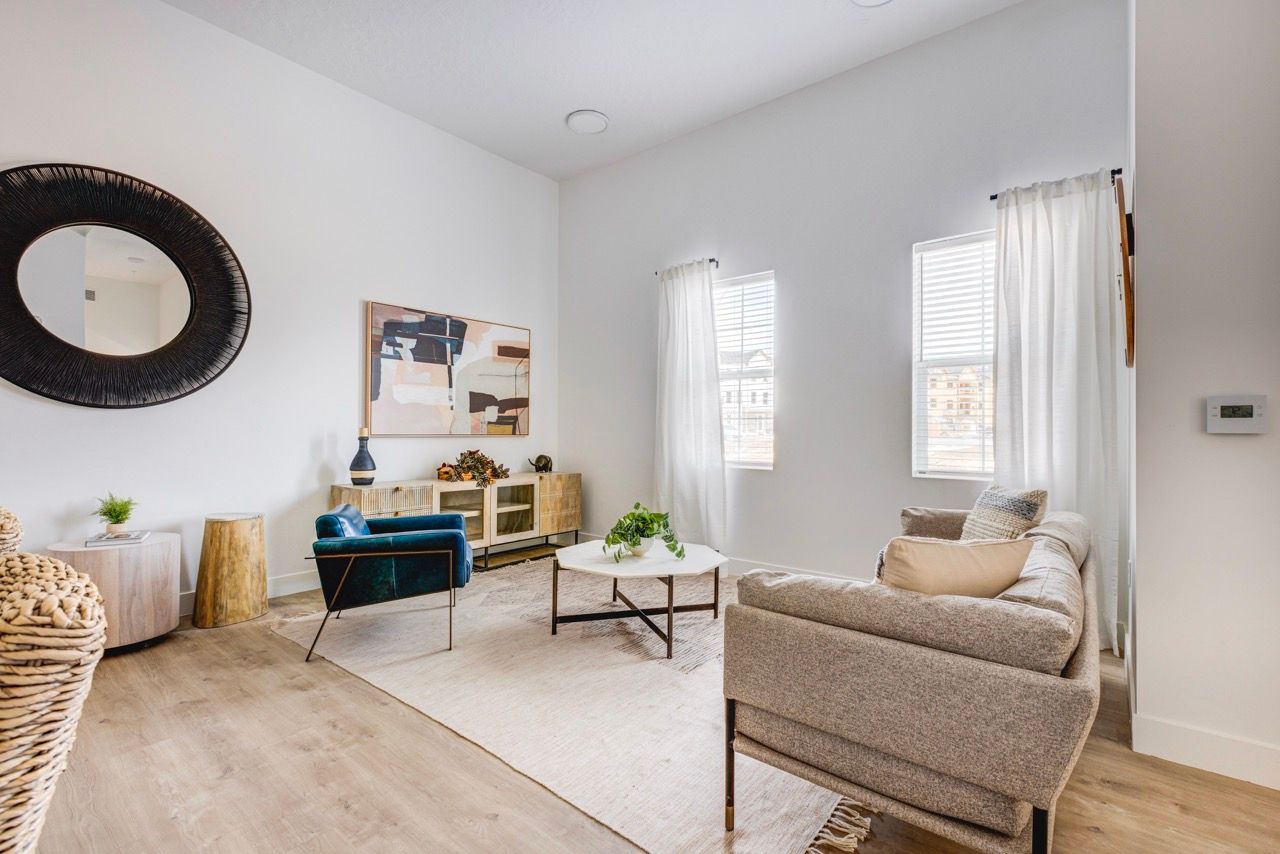 Aria living room with beige sofa, blue accent chair, round woven mirror, and two windows with white curtains.