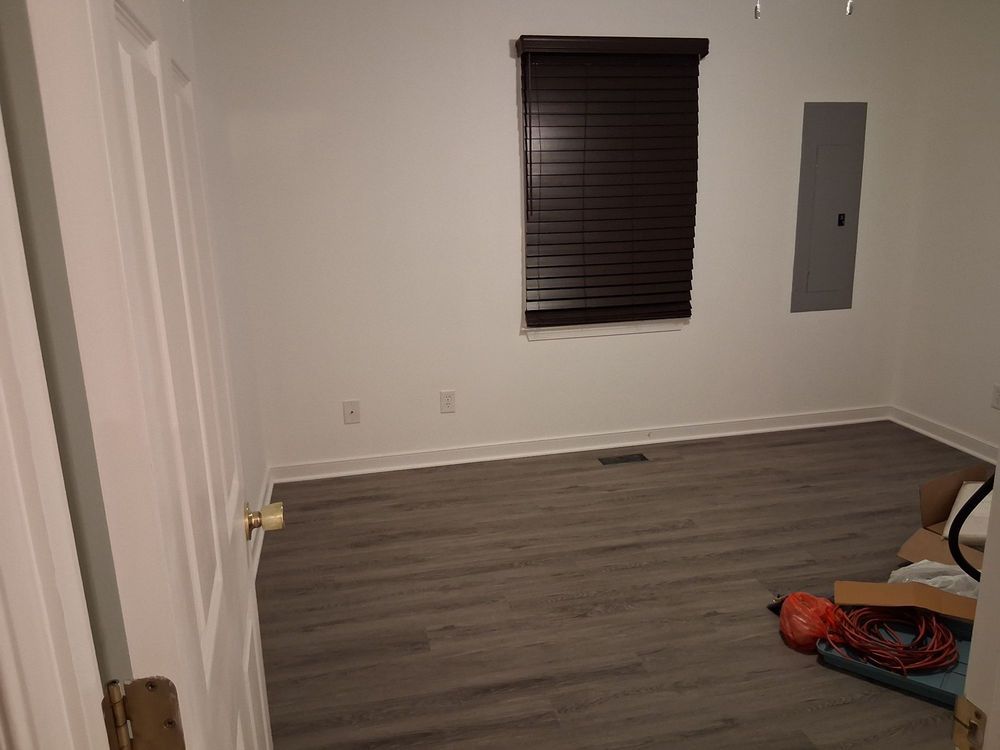 Empty room with gray wood floor, white walls, window blinds, and a wall-mounted electrical panel.