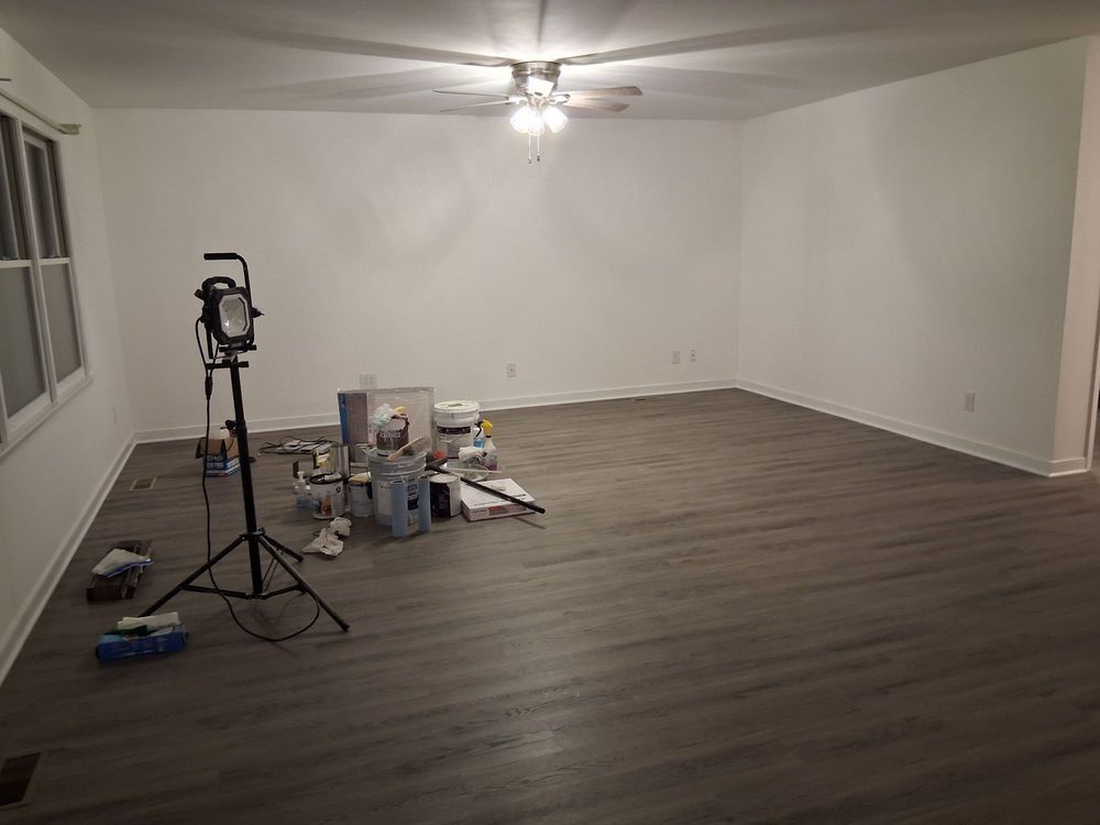 Empty living room with gray wood floors, white walls, and painting supplies near a tripod light stand.