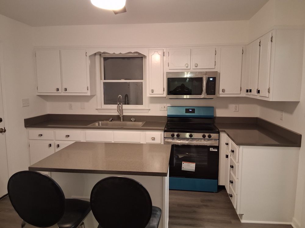 Small white kitchen with dark countertops, central island, stove, sink, and two black chairs.