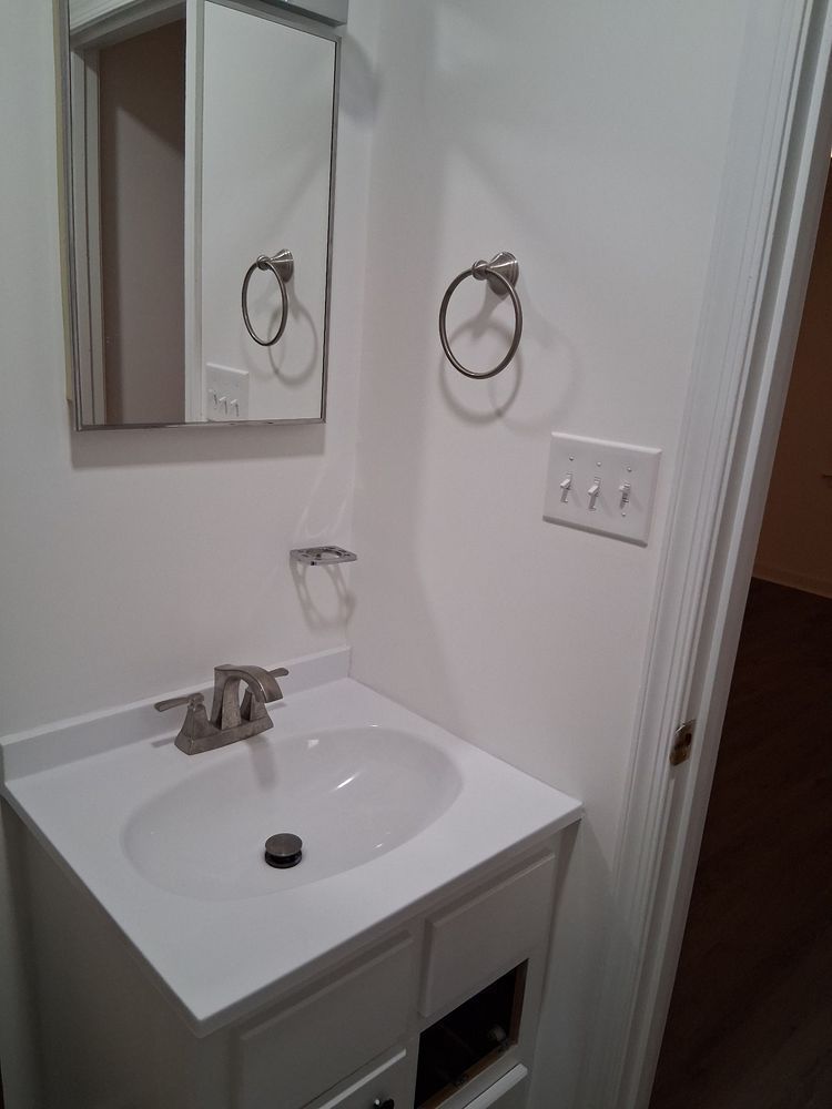 Small bathroom vanity with sink, mirror, towel rings, and light switch on a white wall.