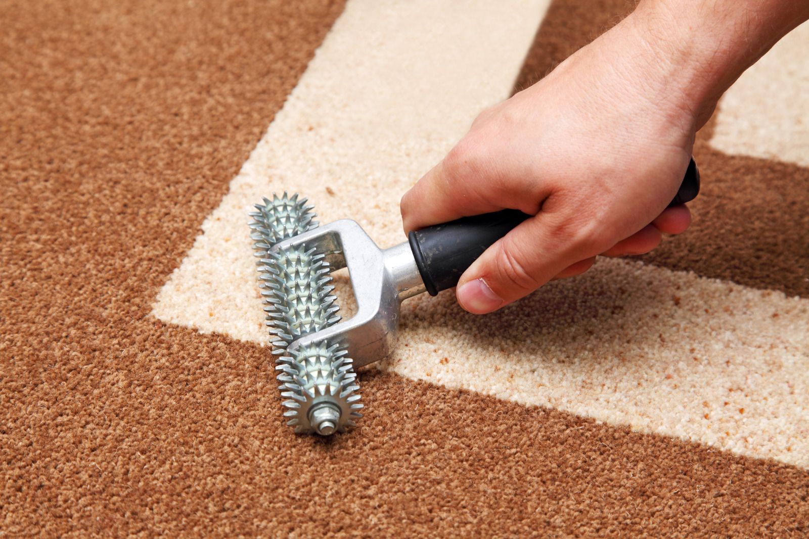 Hand rolling a spiked floor roller over brown carpet near a white taped line