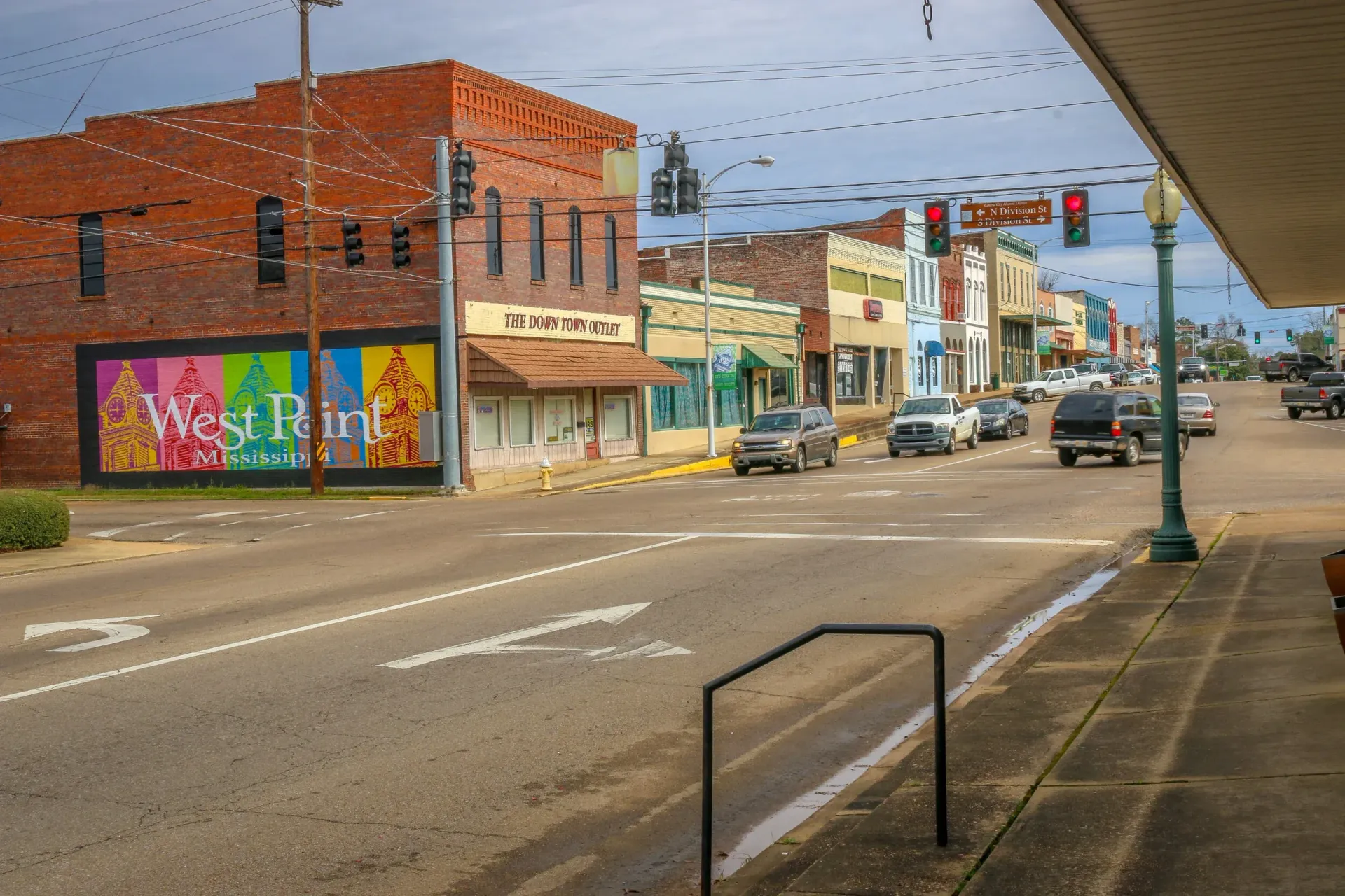 Downtown street with brick storefronts, parked cars, wet pavement, and a bus stop under an overcast sky