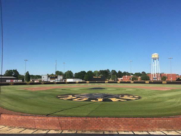 Empty baseball field with green grass, dirt infield, and a water tower under a clear blue sky