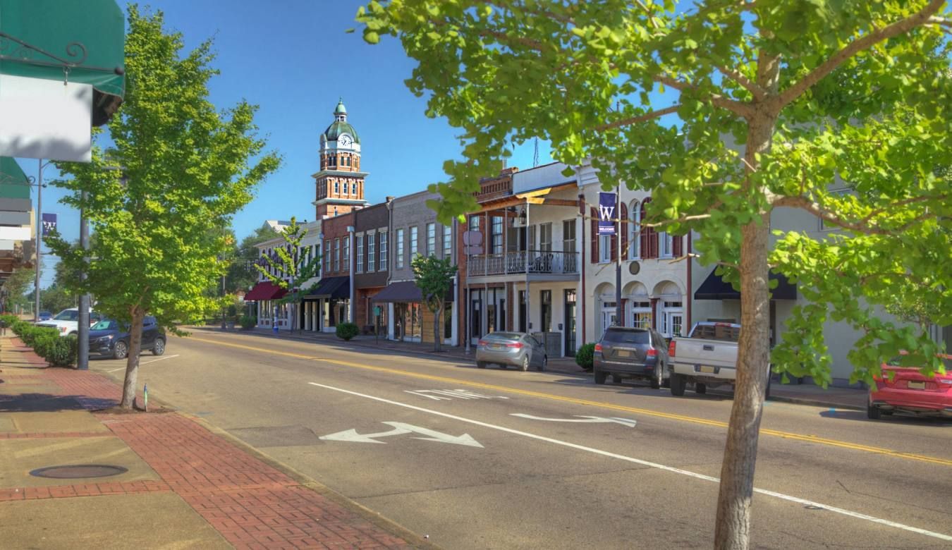 Quiet downtown street with brick buildings, a clock tower, and trees lining the sidewalk on a sunny day
