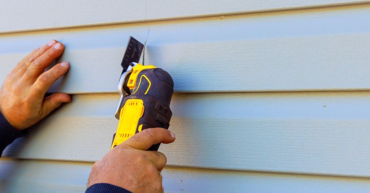 A contractor uses a yellow power tool to cut a damaged blue siding panel while steadying it with one hand.
