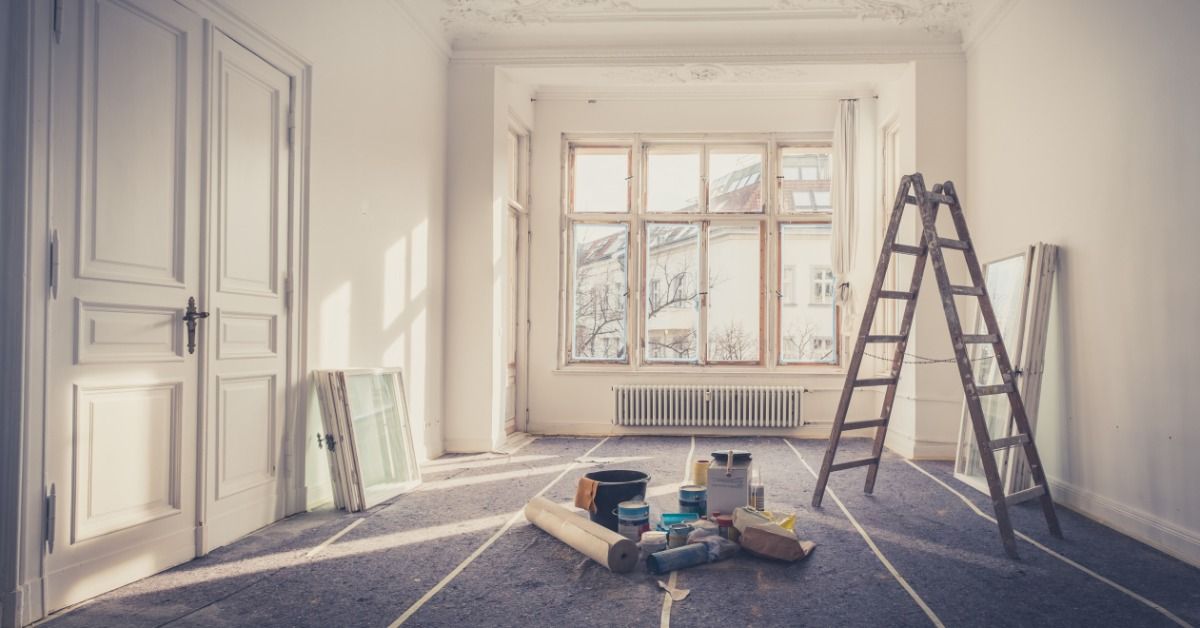 A bright empty room contains a ladder and renovation supplies while protective covering shields the 