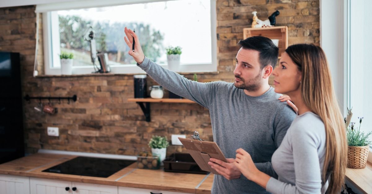 A couple reviews renovation plans in a kitchen as the man points toward a wall and holds a clipboard beside his wife.