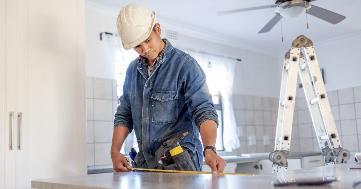 A contractor measures a kitchen countertop with a tape measure as a ladder stands behind him in a bright room.