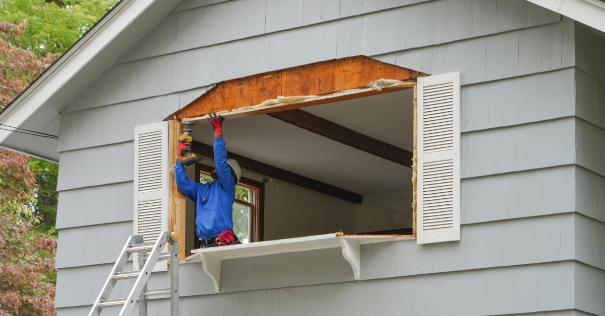Construction worker using tools to remove an old window frame from the exterior of a gray house.