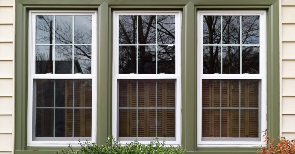 Three white double-hung windows with decorative grid patterns and green trim on the exterior of a beige house.