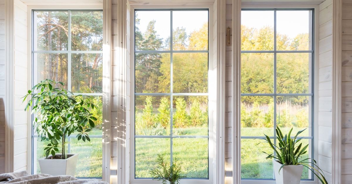 Three tall windows brighten a cozy room with potted plants, pale wood walls, and a view of the green field.