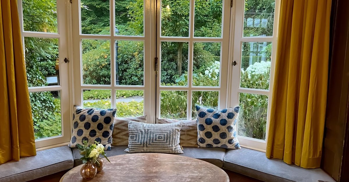 A curved window seat lines tall paneled windows with yellow curtains, patterned pillows, and a garden view.