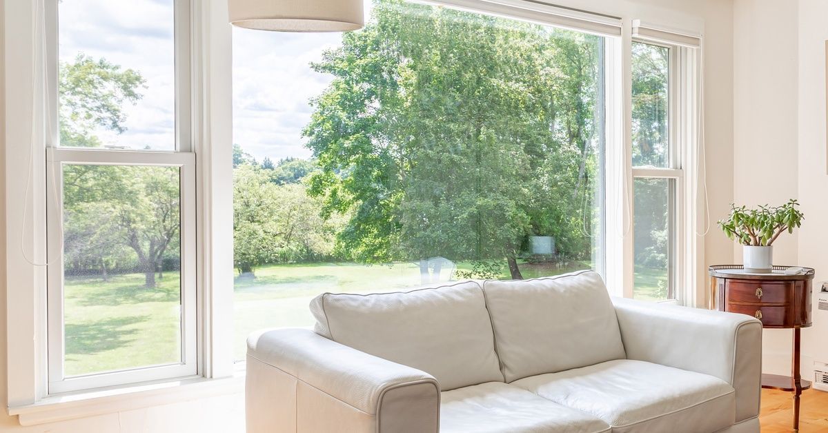 A bright living room features a white sofa before large windows overlooking green trees and a sunny 