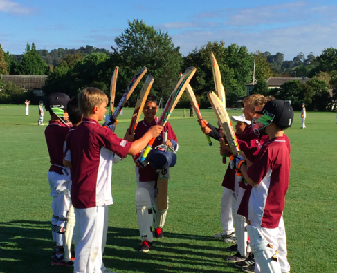 Children in maroon cricket uniforms hold bats aloft, forming a tunnel on a sunny field.