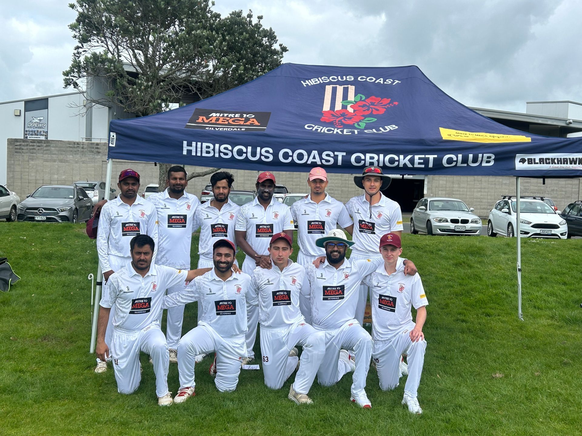 Hibiscus Coast Cricket Club team posing in white uniforms under a blue tent on a grassy field