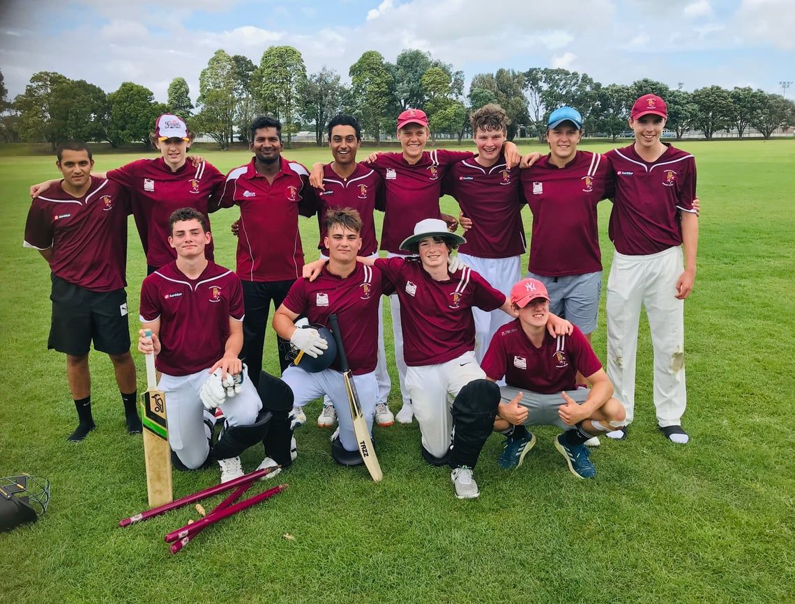 Cricket team in maroon jerseys posing on a grassy field with bats and stumps.