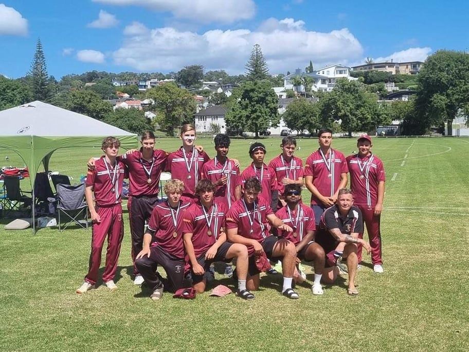 Cricket team in maroon uniforms posing on a grassy field with houses and trees in the background