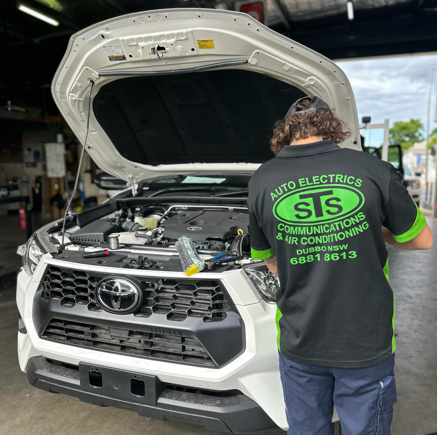 A man wearing a shirt that says auto electrics is working on a car