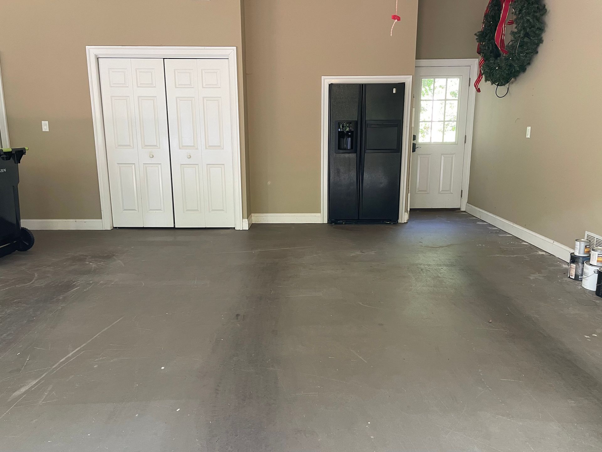 Empty garage with beige walls, white double doors, and a black refrigerator near a side entry door.