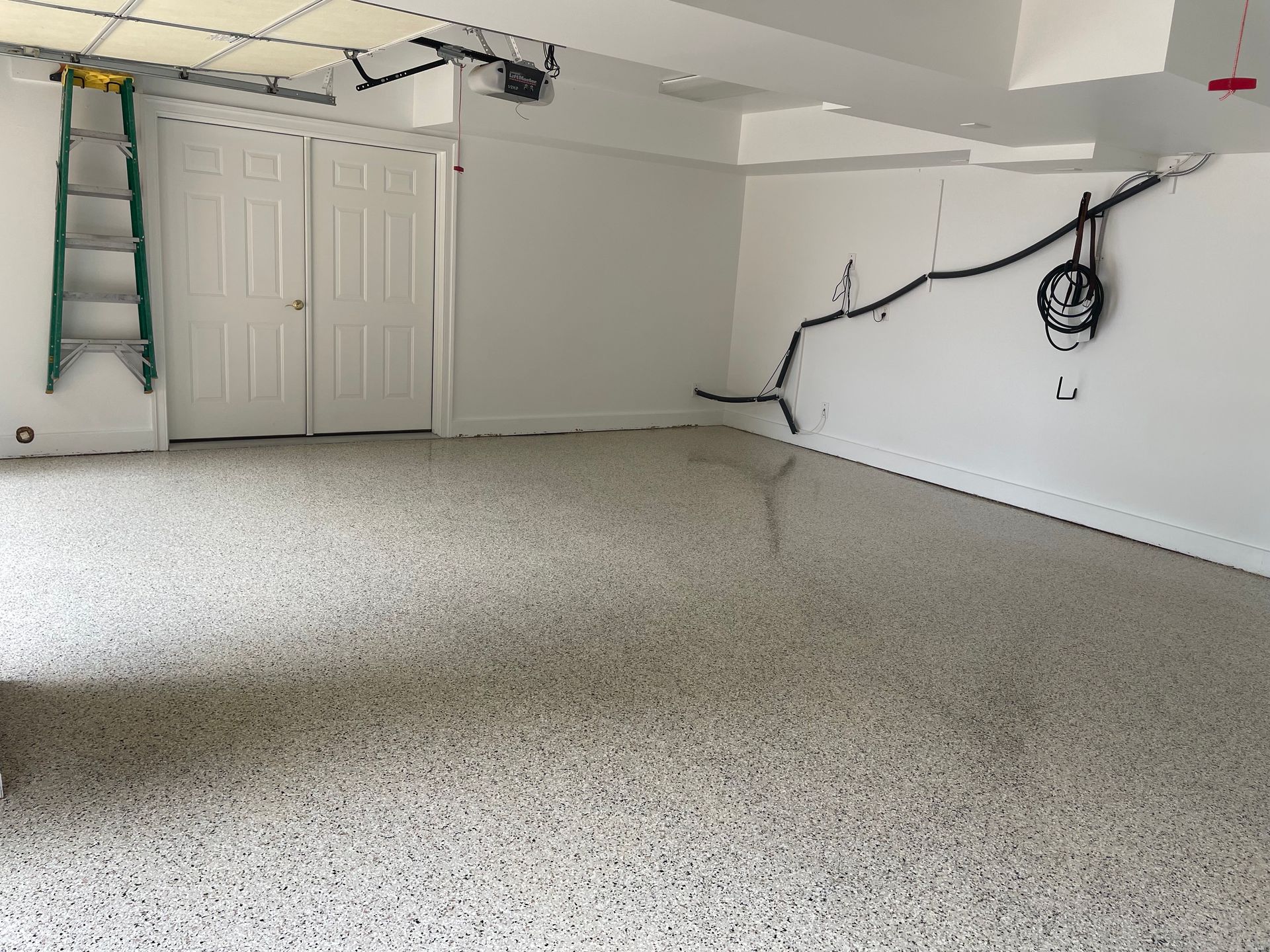 Empty garage with white walls, speckled floor, ladder, and overhead doors.