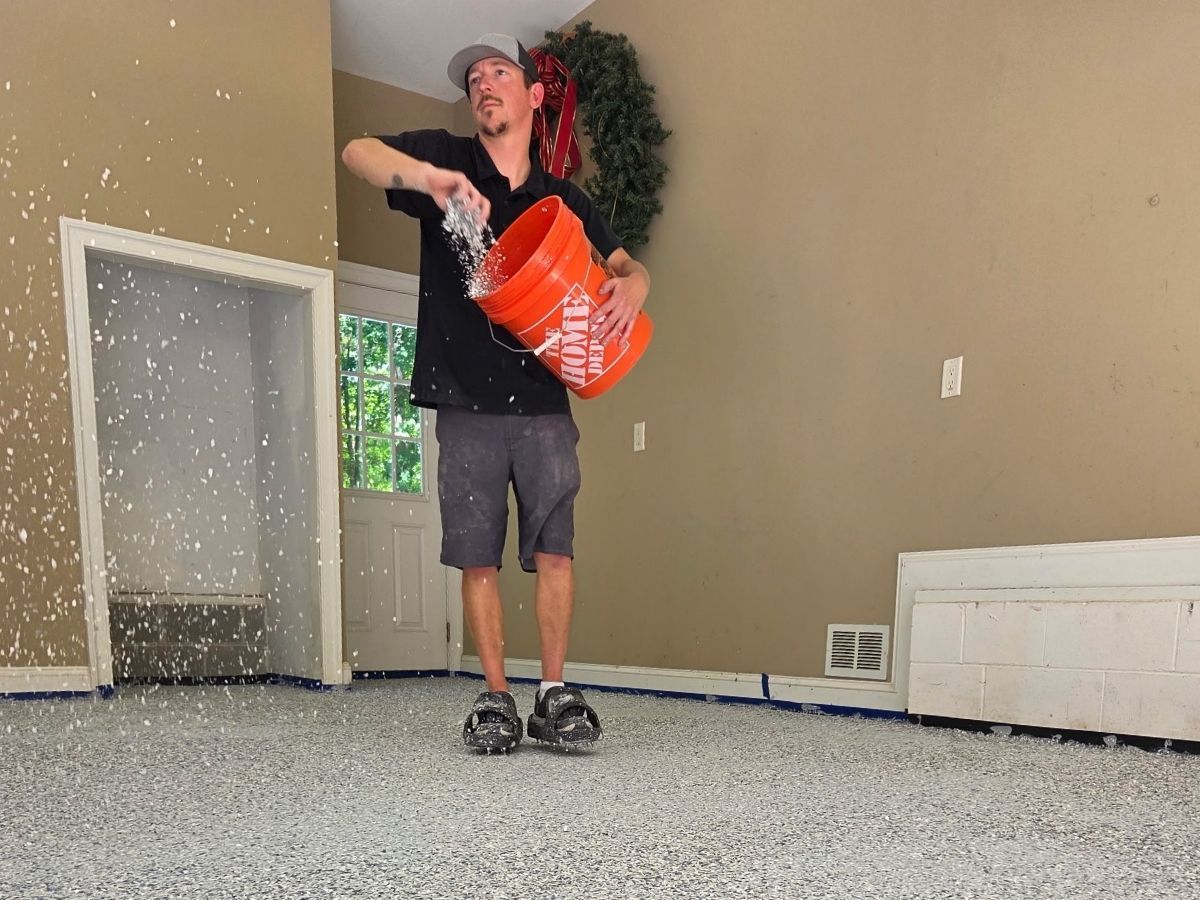 Person sprinkling white confetti from an orange bucket in a room with beige walls and gray carpet