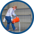 Worker in safety gear pouring liquid from an orange bucket in an industrial facility