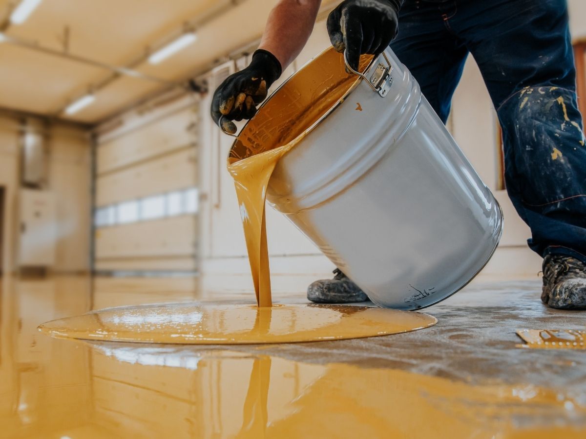 Worker pouring orange paint from a bucket onto a floor in a garage-like space